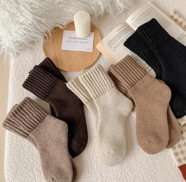 Five pairs of thick ribbed-cuff socks in cream, beige, taupe, brown and black, laid flat on a soft white blanket